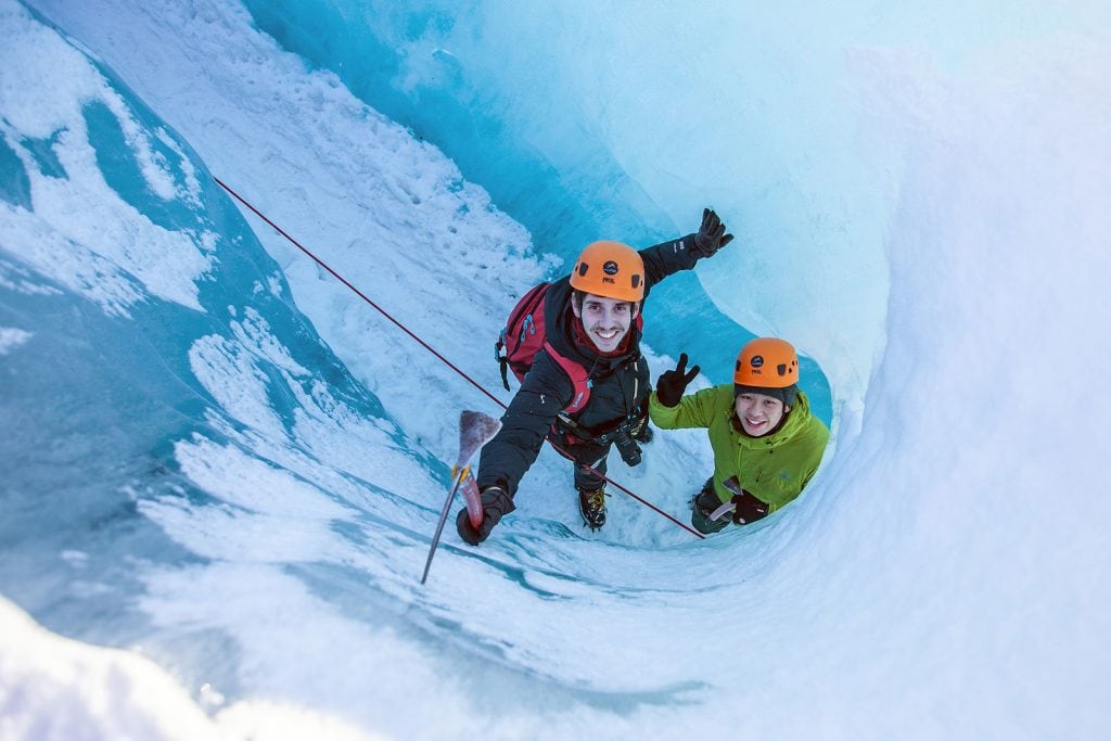 Two climbers, one in pink and one in green jackets, ascend a steep icy slope in Iceland, their faces expressing excitement and determination under their helmets