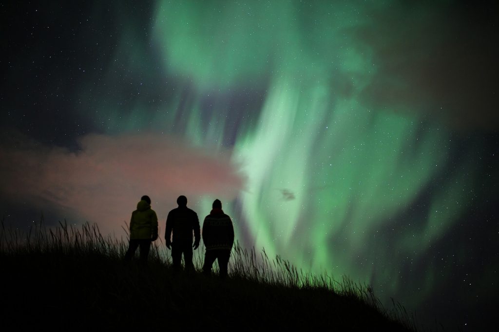 Three people stand silhouetted on a hill, watching the vibrant green and purple hues of the Northern Lights ripple across the night sky in Iceland.