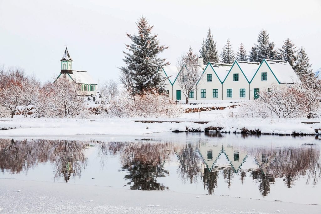 A serene winter scene depicts a quaint village with traditional houses and a church reflected perfectly in the still waters of a nearby lake, all blanketed in snow.