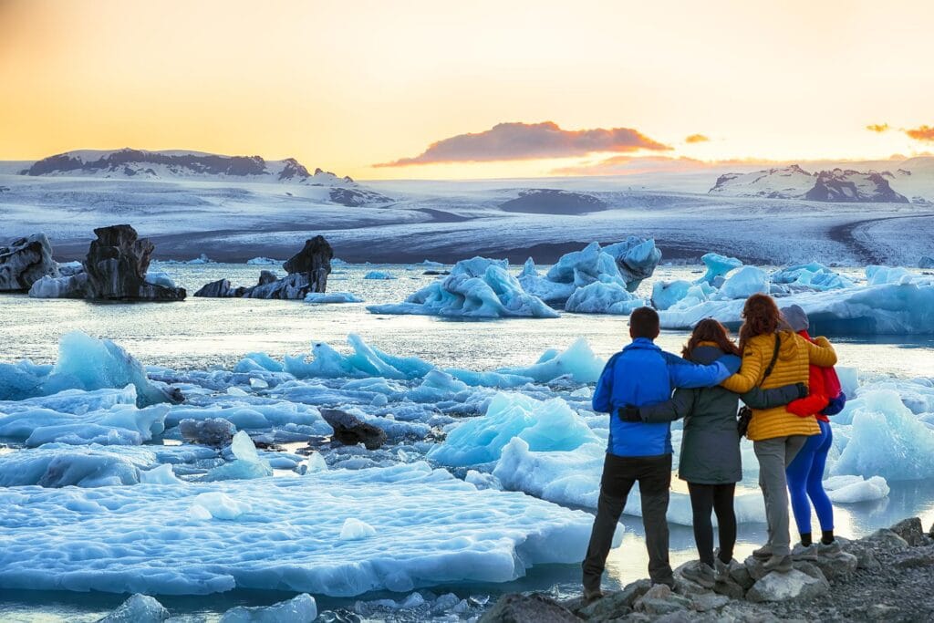 A group of four people stand together, facing a scenic view of a glacial lagoon filled with icebergs at sunset. Snow-covered mountains are visible in the distance.