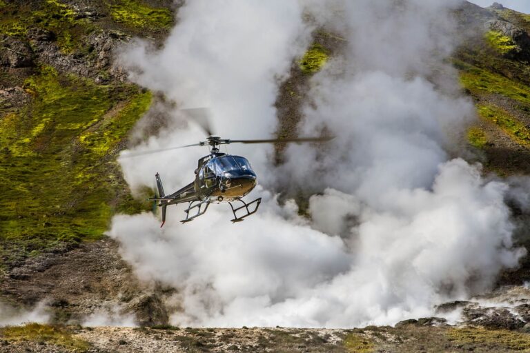 Helicopter flying over a steaming geothermal area in Iceland, showcasing rugged terrain and vibrant moss-covered hills.