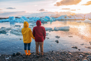A couple in bright rain jackets, one yellow and one red, stand holding hands, looking out over a glacial lagoon with floating icebergs under a cloudy sunset sky.