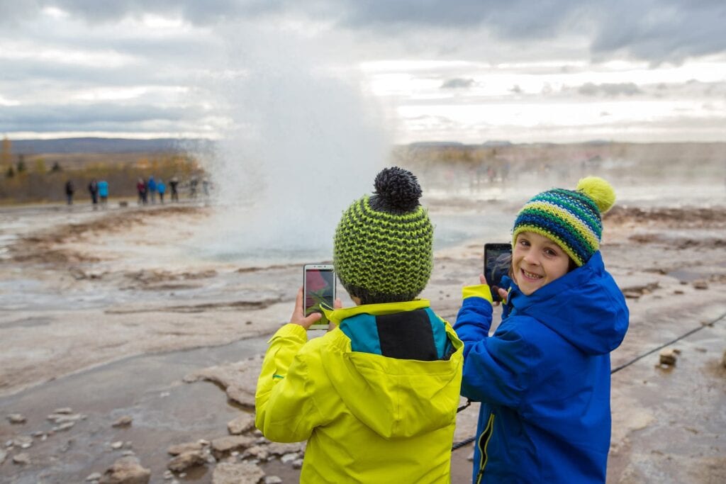 Two children in yellow raincoats stand near a steaming geyser in Iceland.