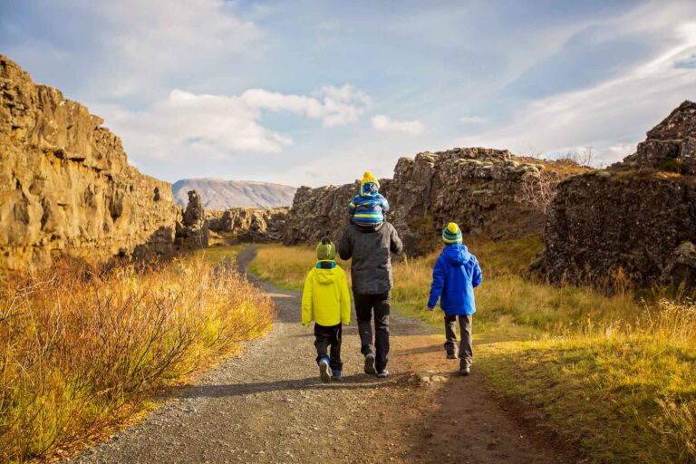 Two children and an adult walk along a gravel path toward a cliffside with rugged Icelandic scenery.