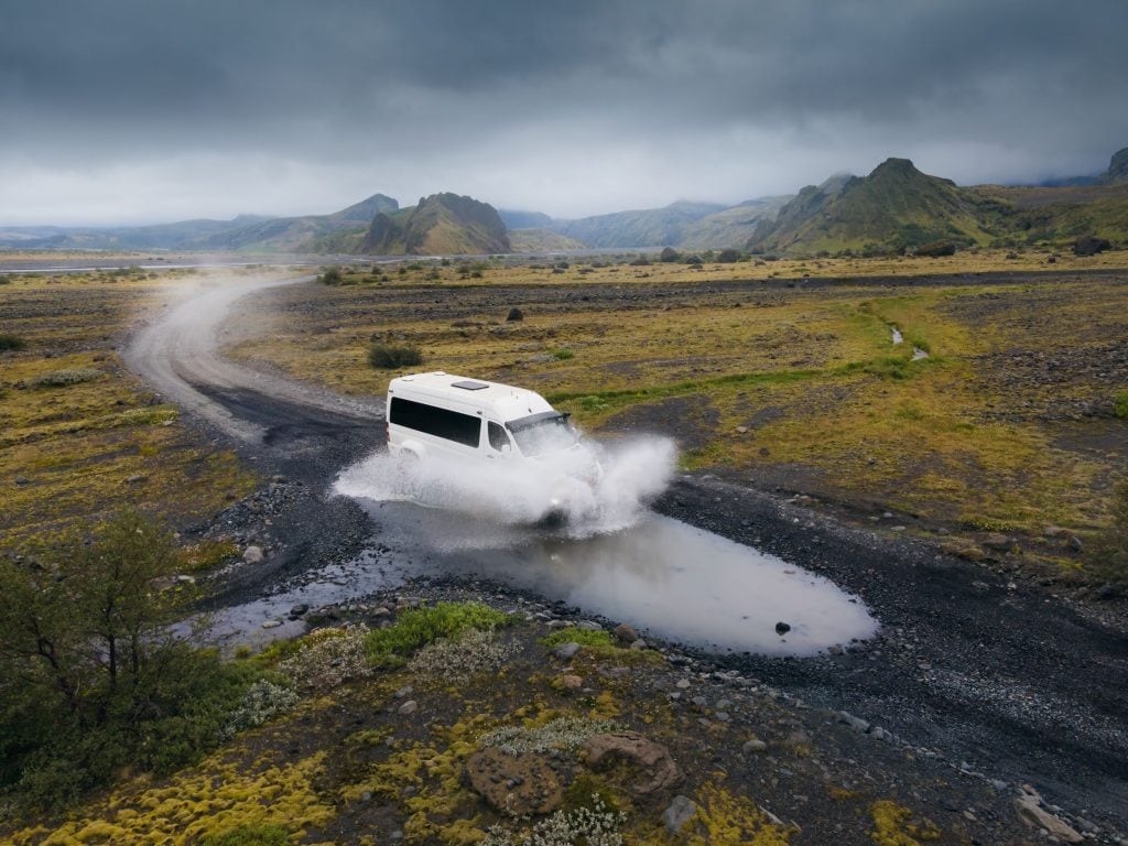 A rugged white jeep fords through a shallow river on a gravel path, splashing water in all directions against the backdrop of Iceland’s dramatic volcanic landscape.