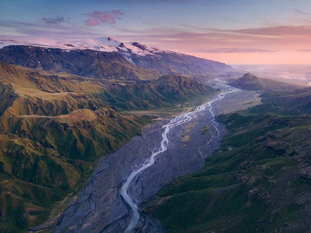 The last light of day casts a warm glow over a sprawling Icelandic valley, with a meandering river cutting through the lush landscape beneath the shadow of a towering glacier.