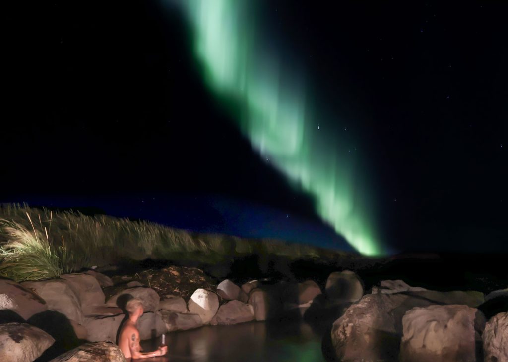 A man is immersed in a hot spring, sipping champagne, with the vibrant Northern Lights dancing above him.