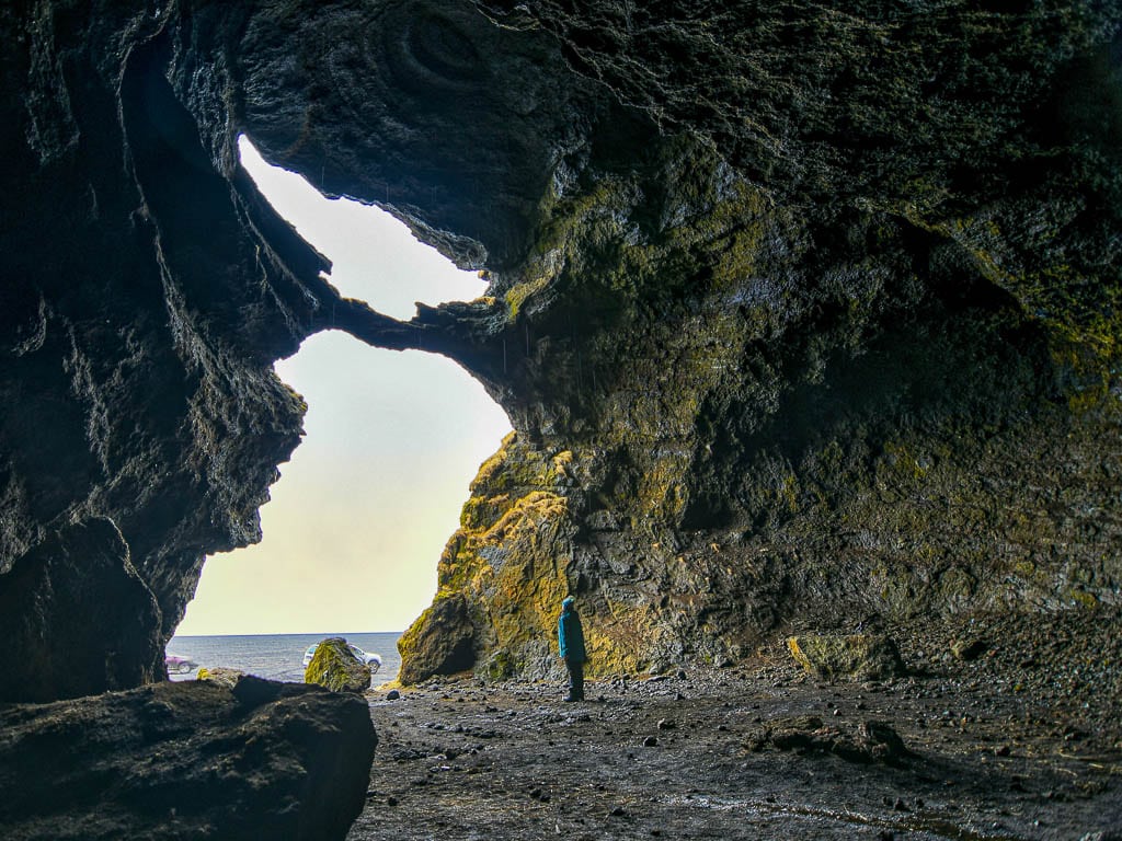 A person standing inside a large cave with an opening that reveals a view of the ocean and sky outside. The cave walls are rugged and covered in moss. ​​