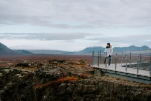 A person in a white jacket stands on a viewing platform, overlooking the rugged landscape of Þingvellir National Park, Iceland.