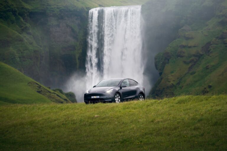 Tesla in Iceland with Waterfall Backdrop: A sleek Tesla car parked on green grass in front of a stunning Icelandic waterfall, highlighting carbon-neutral driving amidst nature.