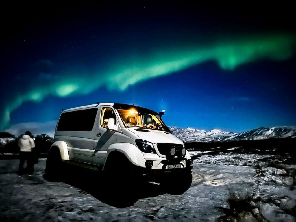 A white off-road van with its headlights on is positioned in a snowy landscape, with the northern lights stretching across the night sky.