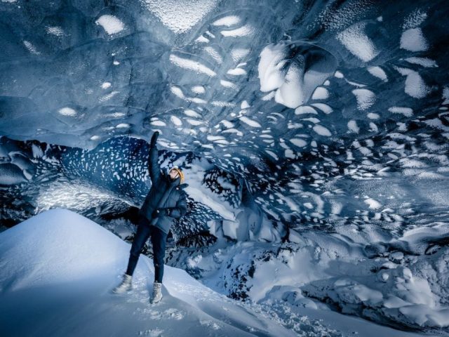 A person with raised arms stands at the entrance of a magnificent ice cave, expressing a sense of triumph or awe. The cave's interior is a frozen wonderland, with snow-covered formations that create an otherworldly landscape.