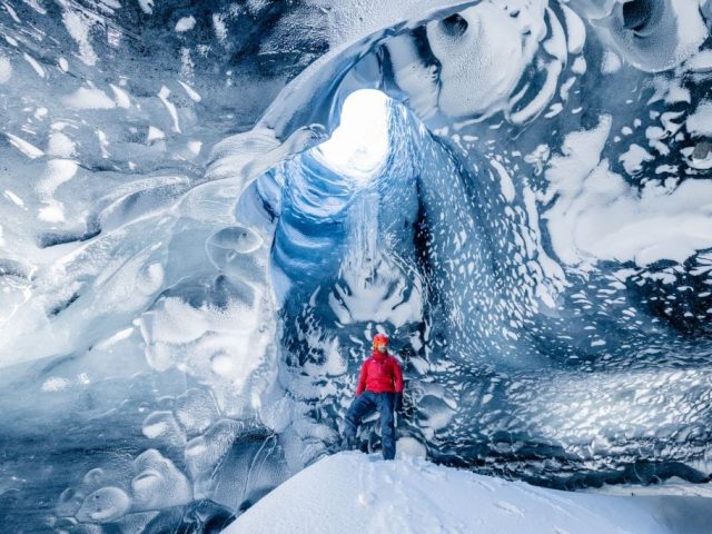 A solo explorer in a red jacket illuminated by natural light inside an ice cave. The cave's ceiling has a skylight where light pours in, creating a surreal and majestic atmosphere around the figure.