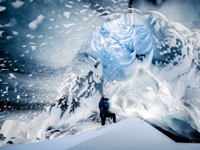 A person standing in front of a large, intricate ice cave with a backdrop of a snow-covered mountain. The cave's walls are textured with layers of snow and ice, resembling a wave above the person, who appears minuscule in comparison.