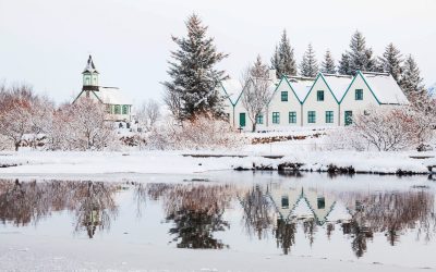 A serene winter scene depicts a quaint village with traditional houses and a church reflected perfectly in the still waters of a nearby lake, all blanketed in snow.
