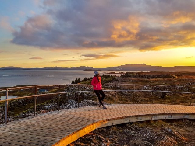 A person in a red jacket stands on a curved path overlooking a scenic sunset at Þingvellir National Park, Iceland.