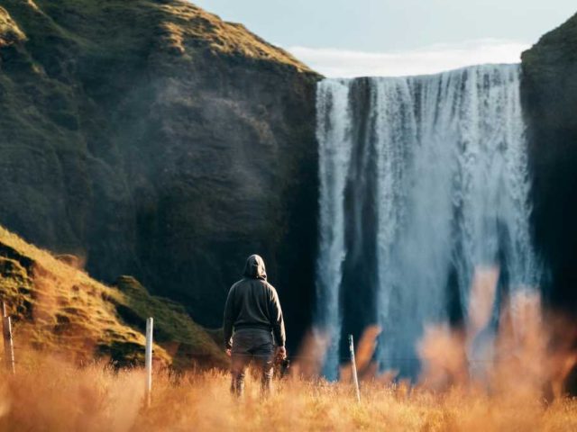 Tourist standing in front of a towering waterfall, observing the powerful cascade from a grassy field.