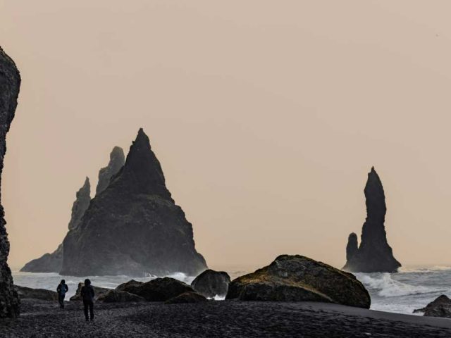 Tall, jagged rock formations rising from the black sand beach, with grasses in the foreground and a soft, hazy sky.