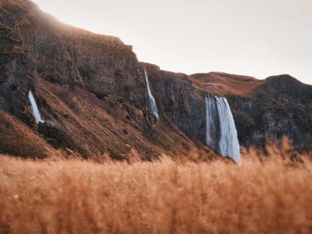 Golden sunset illuminating a waterfall from behind, casting warm hues through the flowing water and mist.