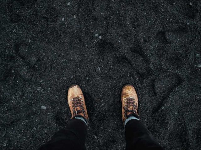 person wearing brown boots standing on a black sand beach, showcasing unique volcanic terrain.