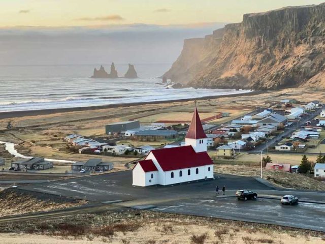 Scenic view of a coastal village with a red-roofed church, surrounded by cliffs and a distant ocean horizon.