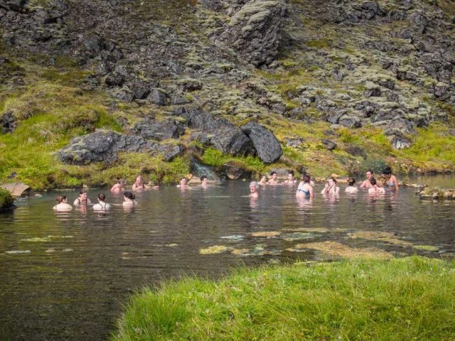 Tourists enjoying a hot spring surrounded by rocky terrain and greenery in the Iceland Highlands during a Landmannalaugar tour.