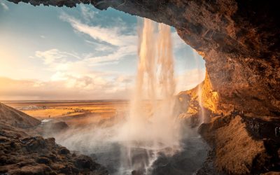 A picturesque view of a waterfall as seen from a cave, with the waterfall cascading down from a rocky overhang and sunlight illuminating the mist created by the falling water. The landscape beyond the waterfall is bathed in a warm, golden light, highlighting the expansive view of the flat plains stretching towards the horizon.