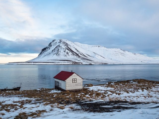 A solitary red-roofed house sits by the shore, with the mountain rising majestically in the background, surrounded by a calm sea and a snow-dusted landscape.
