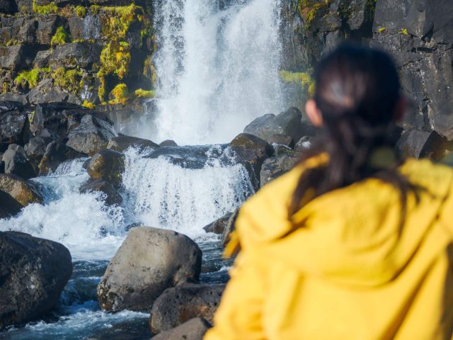 Person in a yellow raincoat observing a waterfall in Thingvellir
