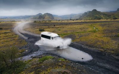 A rugged white jeep fords through a shallow river on a gravel path, splashing water in all directions against the backdrop of Iceland’s dramatic volcanic landscape.