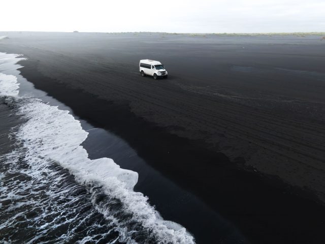 White monster truck cruising along a black sand beach with the sea coastline in view.