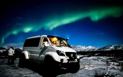 A white off-road van with its headlights on is positioned in a snowy landscape, with the northern lights stretching across the night sky.