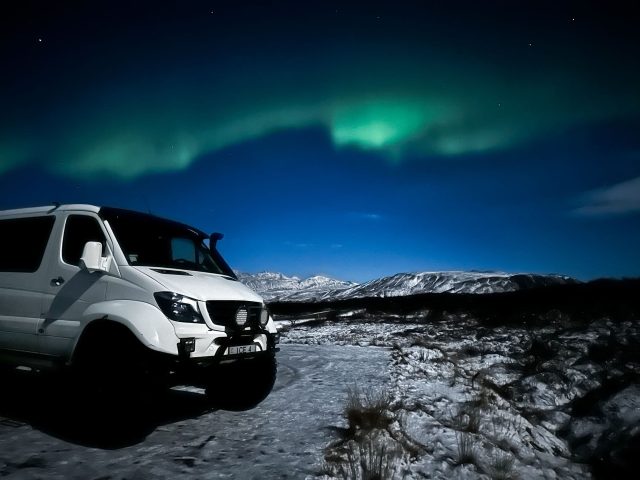 A white off-road van is parked on icy terrain, with the northern lights glowing in green hues above the mountains in the background.