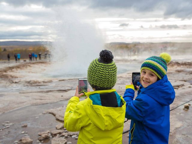 Two children in yellow raincoats stand near a steaming geyser in Iceland.