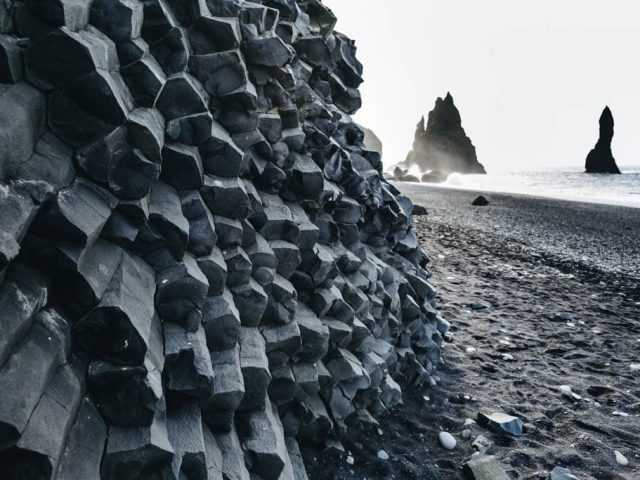A view of a black sand beach featuring distinctive hexagonal basalt columns on the left, with a hazy sea stack visible in the background amidst a misty ocean spray. The scene captures the unique geological formations typical of volcanic landscapes.