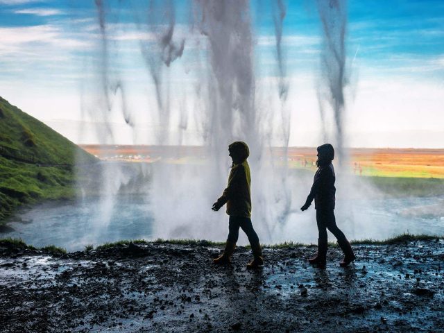 Two people walk behind the cascading water of Seljalandsfoss waterfall in Iceland, with sunlight and a rainbow visible in the distance.