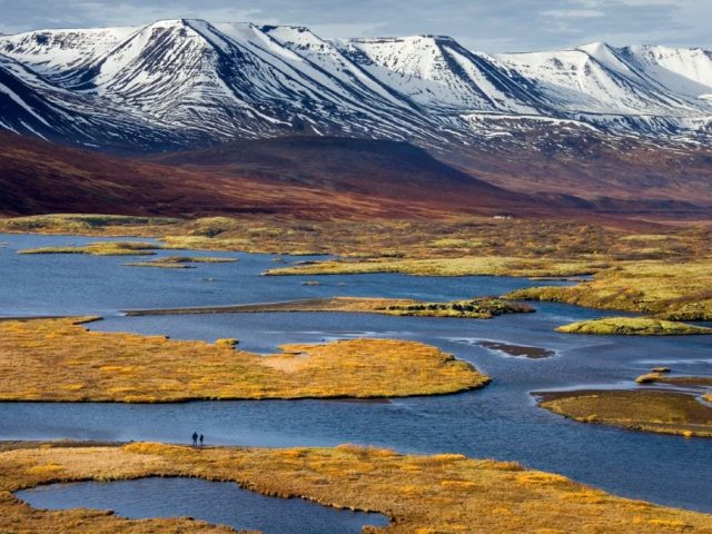 An expansive view of a marshy landscape with winding waterways, golden-hued grasses, and a solitary figure standing on a narrow land strip, set against a backdrop of distant snow-capped mountains under a clear sky.
